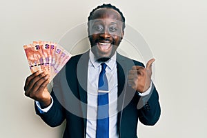 Handsome young black man holding south african rand banknotes pointing thumb up to the side smiling happy with open mouth
