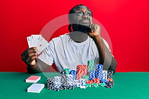 Handsome young black man playing poker holding cards serious face thinking about question with hand on chin, thoughtful about