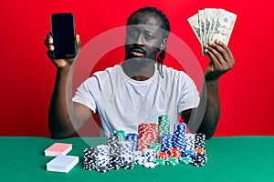 Handsome young black man playing poker holding smartphone and dollars clueless and confused expression