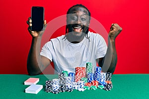 Handsome young black man playing poker holding smartphone screaming proud, celebrating victory and success very excited with