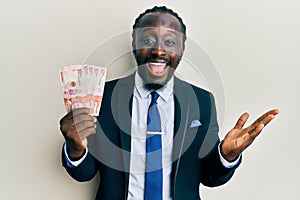 Handsome young black man wearing business suit holding 10 colombian pesos celebrating achievement with happy smile and winner