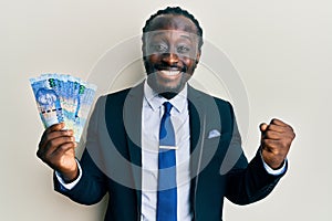 Handsome young black man wearing business suit holding 100 rands banknotes screaming proud, celebrating victory and success very