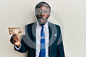Handsome young black man wearing business suit holding 1000 yen banknotes scared and amazed with open mouth for surprise,