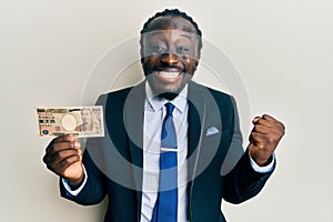 Handsome young black man wearing business suit holding 1000 yen banknotes screaming proud, celebrating victory and success very