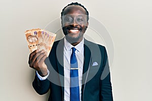 Handsome young black man wearing business suit holding 20 rands banknotes looking positive and happy standing and smiling with a