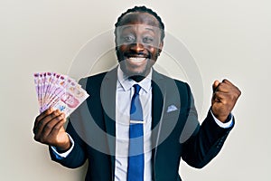 Handsome young black man wearing business suit holding mexican pesos banknotes screaming proud, celebrating victory and success