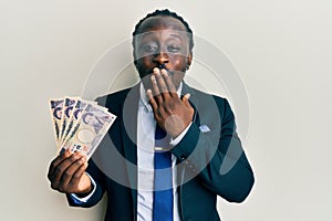 Handsome young black man wearing business suit holding yens banknotes covering mouth with hand, shocked and afraid for mistake