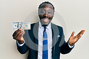 Handsome young black man wearing business suit and tie holding 100 dollars celebrating achievement with happy smile and winner