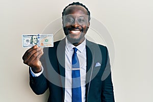 Handsome young black man wearing business suit and tie holding 100 dollars looking positive and happy standing and smiling with a
