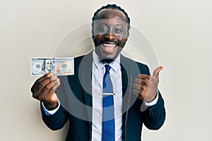 Handsome young black man wearing business suit and tie holding 100 dollars pointing thumb up to the side smiling happy with open