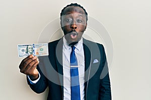 Handsome young black man wearing business suit and tie holding 100 dollars scared and amazed with open mouth for surprise,