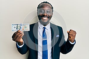 Handsome young black man wearing business suit and tie holding 100 dollars screaming proud, celebrating victory and success very