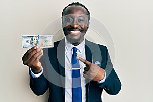 Handsome young black man wearing business suit and tie holding 100 dollars smiling happy pointing with hand and finger