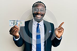 Handsome young black man wearing business suit and tie holding 100 dollars smiling happy pointing with hand and finger to the side