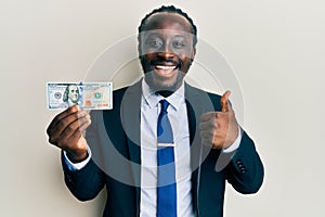 Handsome young black man wearing business suit and tie holding 100 dollars smiling happy and positive, thumb up doing excellent