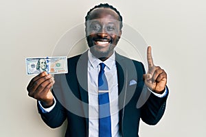 Handsome young black man wearing business suit and tie holding 100 dollars smiling with an idea or question pointing finger with