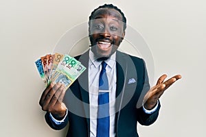 Handsome young black man wearing business suit and tie holding australian dollars celebrating achievement with happy smile and