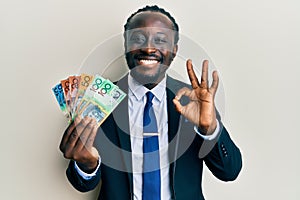 Handsome young black man wearing business suit and tie holding australian dollars doing ok sign with fingers, smiling friendly