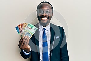 Handsome young black man wearing business suit and tie holding australian dollars looking positive and happy standing and smiling