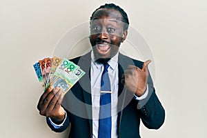 Handsome young black man wearing business suit and tie holding australian dollars pointing thumb up to the side smiling happy with