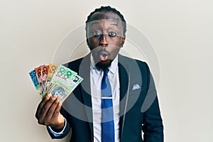 Handsome young black man wearing business suit and tie holding australian dollars scared and amazed with open mouth for surprise,