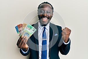 Handsome young black man wearing business suit and tie holding australian dollars screaming proud, celebrating victory and success