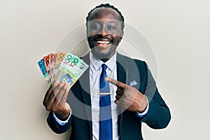 Handsome young black man wearing business suit and tie holding australian dollars smiling happy pointing with hand and finger