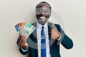 Handsome young black man wearing business suit and tie holding australian dollars smiling happy and positive, thumb up doing