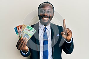 Handsome young black man wearing business suit and tie holding australian dollars smiling with an idea or question pointing finger