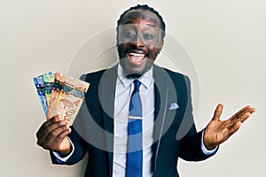 Handsome young black man wearing business suit and tie holding canadian dollars celebrating achievement with happy smile and
