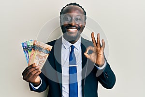 Handsome young black man wearing business suit and tie holding canadian dollars doing ok sign with fingers, smiling friendly