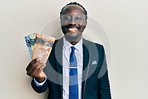 Handsome young black man wearing business suit and tie holding canadian dollars looking positive and happy standing and smiling