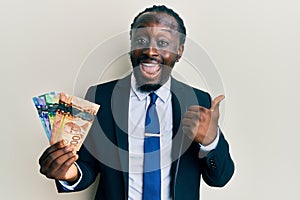 Handsome young black man wearing business suit and tie holding canadian dollars pointing thumb up to the side smiling happy with