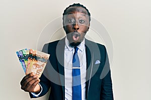 Handsome young black man wearing business suit and tie holding canadian dollars scared and amazed with open mouth for surprise,