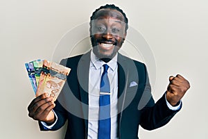 Handsome young black man wearing business suit and tie holding canadian dollars screaming proud, celebrating victory and success