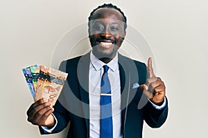 Handsome young black man wearing business suit and tie holding canadian dollars smiling with an idea or question pointing finger