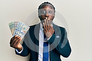 Handsome young black man wearing business suit and tie holding dollars covering mouth with hand, shocked and afraid for mistake