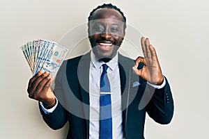 Handsome young black man wearing business suit and tie holding dollars doing ok sign with fingers, smiling friendly gesturing