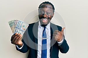 Handsome young black man wearing business suit and tie holding dollars pointing thumb up to the side smiling happy with open mouth