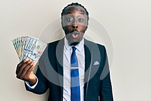 Handsome young black man wearing business suit and tie holding dollars scared and amazed with open mouth for surprise, disbelief