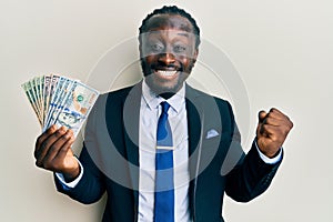 Handsome young black man wearing business suit and tie holding dollars screaming proud, celebrating victory and success very