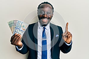 Handsome young black man wearing business suit and tie holding dollars smiling with an idea or question pointing finger with happy