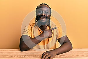 Handsome young black man wearing casual clothes sitting on the table cheerful with a smile of face pointing with hand and finger