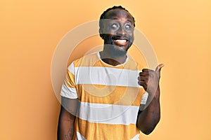 Handsome young black man wearing casual yellow tshirt smiling with happy face looking and pointing to the side with thumb up