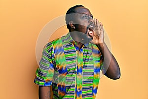 Handsome young black man wearing colorful ethnic clothes shouting and screaming loud to side with hand on mouth