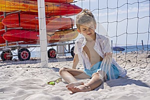 Handsome young boy in blue shorts and white shirt sits on the white sand on the beach. Plays with white sand.