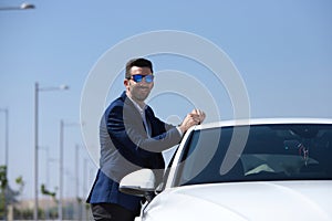 Handsome young businessman with beard and blue suit leaning on his white sports car. The man is wearing sunglasses. Business
