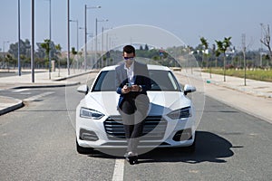 Handsome young businessman with beard and blue suit standing next to his white sports car. The man is wearing sunglasses and