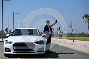 Handsome young businessman with beard and blue suit standing next to his white sports car. The man wears sunglasses and takes