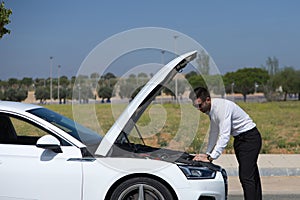 Handsome young businessman with beard looks at the engine of his car which is broken down on the road. The man is in a hurry